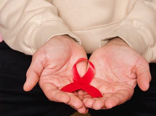 Foto de mãos estendidas em forma de conhca de um homem com calça preta e blusa creme. Sob as mãos do homem está um laço vermelho que representa a luta pela hemofilia. A imagem ilustra o artigo "Hemofilia na Europa: História, Causas Genéticas e Psicanalíticas" do blog do Homero Mônaco para o site Online Psicanálise.