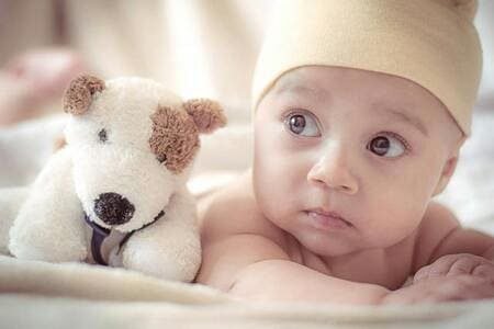 Criança de aproximadamente 1 ano deitada de bruços, com gorro amarelo claro, e um cachorro de pelúcia branco e marrom ao seu lado, representando o seu objeto transacional. A foto ilustra o artigo "Donald Winnicott e a teoria sobre o objeto transicional" escrito pelo psicanalista Homero Monaco para o blog do site Online Psicanalista.