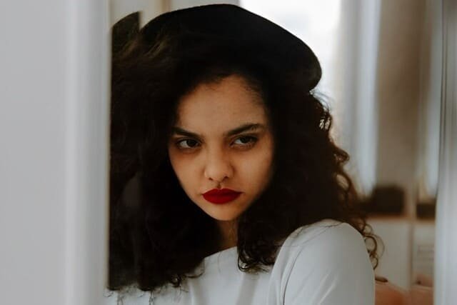 Foto de menina co cabelos cacheados castanho escuros, com boina preta e camiseta branca, usando batom vermelho e com cara de inveja. A foto ilustra o artigo "O Poder Destrutivo da Inveja Revelado" escrito pelo psicanalista Homero Mônaco para o site Online Psicanalista.
