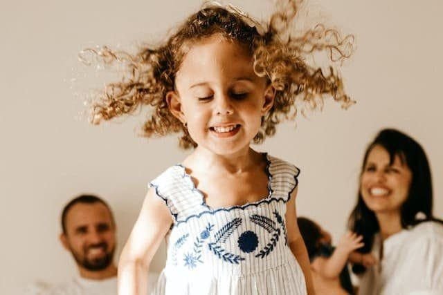 Foto de uma menina de cabelo castanho claro enrolado em primeiro plano, pulando na cama. Em segundo plano aparece o pai, ao lado da mãe com um bebê no colo. Ambos estão sorrindo. A foto ilustra o artigo "Revelando o poder das experiências da infância" escrito pelo psicanalista Homero Mônaco para o site Online Psicanalista.