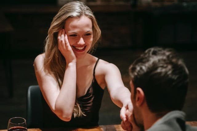 Foto de uma mulher loira, com vestido preto, sentada de frente para a câmera. A sua frente está um homem beijando a sua mão. Ambos estão sentados em uma mes de restaurante. A foto ilustra o artigo "Revigore o seu relacionamento com estas estratégias comprovadas" escrito pelo psicanalista Homero Mônaco para o site online psicanalista.