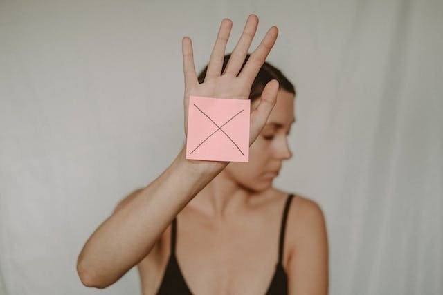 Foto de frente de uma mulher branca, com regata preta, virando o rosto para o lado direito e estendendo a mão para a frente. Na palma de sua mão há um post-it rosa com um "X". A foto ilustra o artigo "Sinais de Alerta no Relacionamento: Não os ignore!" escrito pelo psicanalista Homero Monaco para o site online psicanalista.