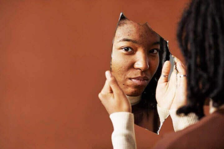 Foto do rosto de uma adolescente negra refletida em um espelho quebrado que ela segura em suas mãos. A foto ilustra o artigo 10 Melhores Maneiras como a Psicanálise Impulsiona a Autoidentidade escrito pelo psicanalista Homero Mônaco para o site Online Psicanalista.