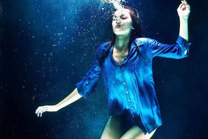 Foto de uma mulher com camisa azul, mergulhada em uma piscina, de olho fechado, soltando bolinhas de ar pela boca como se estivesse afundando e se afogando. A foto ilustra o artigo Como a Psicanálise trata a depressão escrito no blog do psicanalista Homero Mônaco para o site A Sessão de Terapia.