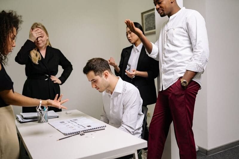 Foto de um homem sentado na mesa do escritório, de cabeça baixa, submisso, enquanto outras pessoas em pé a sua volta parecem estar lhe dando bronca e humilhando. A imagem ilustra o artigo Masoquismo primário e masoquismo secundário uma exploraçãp detalhada escrito pelo Psicanalista Homero Mônaco para site Online Psicanalista.