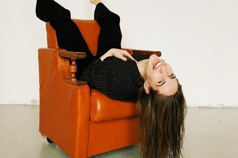 Foto de uma mulher, sentada invertida em uma poltrona de couro caramelo. Seus pés estão no encosto da poltrona, seu corpo apoiado no assento dela, e sua cabeça com seu longo cabelo, estão para fora da cadeira. A mulher olha para a câmera, como se estivesse feliz com a sua recuperação emocional conseguida no setting de terapia. A foto ilustra o artigo O poder da Terapia pa a sua Recuperação Emocional escrito pelo psicanalista Homero Mônaco para o site Online Psicanalista.