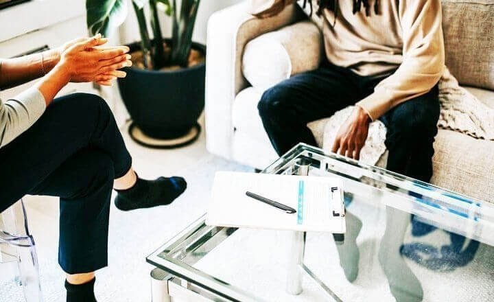 Foto de um homem negro sentado em um sofá bege, enquanto conversa com um terapeuta sentado à sua frente. A foto ilustra o artigo 10 Melhores Maneiras para Construir a Resiliência Emocional com a Psicanálise escrito pelo psicanalista Homero Mônaco para o site Online Psicanalista.
