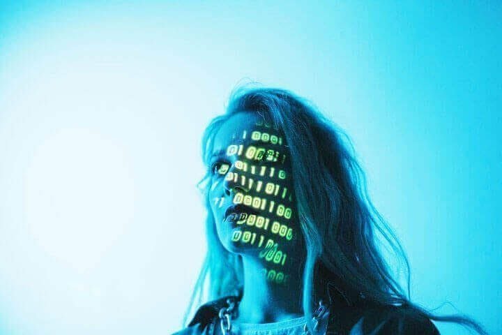 Foto do rosto de uma mulher loira, com parede branca ao fundo, e na sua cara aparecem códigos de programação como se ela estivesse visualizando algo.