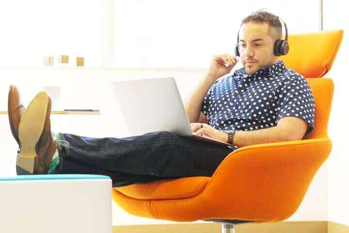 Foto de um homem sentado em uma cadeira laranja, em um escritório, com fone de ouvidos e olhando para o laptop que está em cima das suas pernas. Ele está com as pernas erguidas, com os pés em cima de uma mesa e com roupa informal, representando o equilíbrio entre o trabalho e a vida pessoal. A foto ilustra o artigo 5 Estratégias para Equilibrar Trabalho e Crescimento Pessoal escrito pelo psicanalista Homero Mônaco para o site Online Psicanalista.
