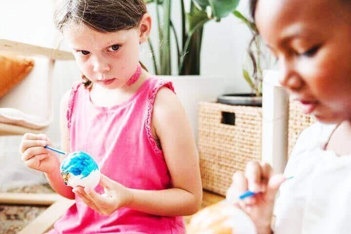 Foto de uma menina sentada em um sofá pintando um ovo que está na sua mão, enquanto olha para outra menina sentada no mesmo sofá, também pintando um ovo. A primeira menina parece estar olhando para aprender com a outra, mas existe um certo olhar de inveja em sua face. A foto ilustra o artigo 75% do seu Comportamento foi Moldado pelas Experiências da sua Infância escrito pelo psicanalista Homero Mônaco para o site Online Psicanalista.
