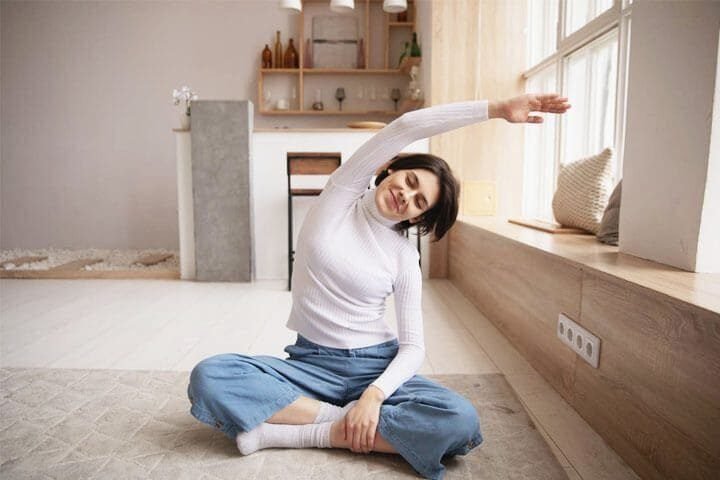 Foto de uma mulher de cabelos castanhos sentada no chão estirando-se com cara de positivismo. A foto ilustra o artigo 7 Maneiras como a terapia ajuda a superar hábitos autossabotadores escrito pelo psicanalista Homero Mônaco para o site Online Psicanalista.
