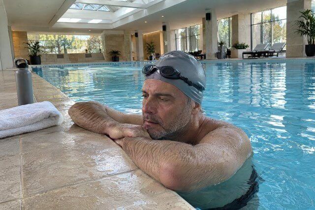 Homem apoiado na borda de uma piscina logo depois de nadar, com os óculos de natação na testa e os braços apoiados na borda enquanto recupera o fôlego. A expressão demonstra cansaço físico moderado, e a postura sugere uma pausa após a atividade. A cena retrata o exercício como parte de uma rotina de cuidado pessoal e bem-estar psicológico. Imagem utilizada para ilustrar o artigo “10 Estratégias para Promover o Bem-estar Psicológico” escrito por Homero Mônaco Psicanalista especializado em neurociências e comportamento.