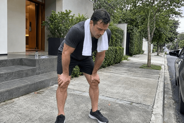 Homem de cerca de 40 anos na calçada diante de casa, curvado para a frente com as mãos apoiadas nos joelhos depois de uma corrida matinal. Uma toalha está pendurada no pescoço, e o rosto demonstra cansaço enquanto recupera o fôlego. A cena transmite esforço, recuperação e a capacidade de continuar mesmo depois de um momento exigente. Imagem utilizada para ilustrar o artigo “10 Melhores Maneiras para Construir a Resiliência Emocional com a Psicanálise” escrito por Homero Mônaco Psicanalista especializado em neurociências e comportamento.