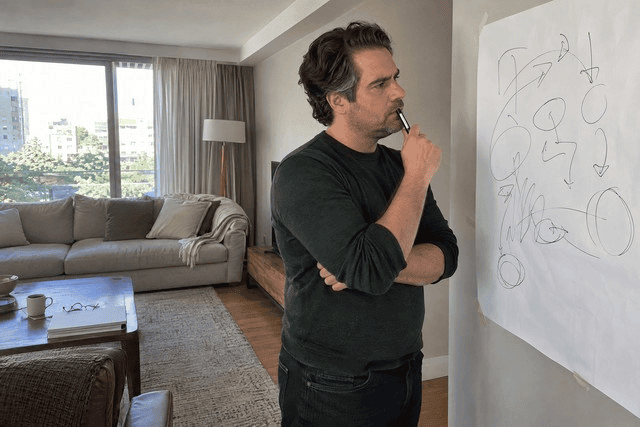 Homem de cerca de 40 anos em pé diante de uma folha grande de papel presa à parede, segurando uma caneta perto da boca enquanto observa rabiscos e desenhos abstratos. O ambiente é uma sala pequena de casa, com poucos objetos de trabalho. Sua postura concentrada sugere uma pausa para organizar ideias e encontrar uma solução para um problema complexo. Imagem utilizada para ilustrar o artigo “10 Técnicas Criativas para o Sucesso na Resolução de Problemas” escrito por Homero Mônaco Psicanalista especializado em neurociências e comportamento.