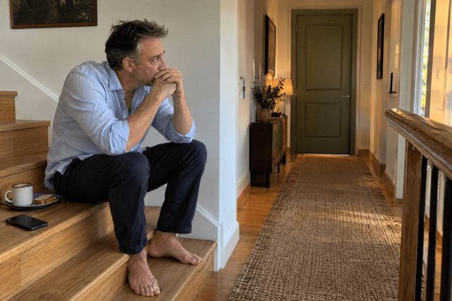 Homem de cerca de 47 anos sentado no meio da escada de madeira de sua casa, com os cotovelos apoiados nos joelhos e as mãos entrelaçadas diante da boca, olhando para a porta fechada de um quarto no corredor. Uma xícara de chá intocada e um prato com fatias de pão estão ao seu lado. Sua postura curvada e expressão cansada transmitem preocupação e impotência diante da dificuldade de ajudar alguém querido. Imagem utilizada para ilustrar o artigo “7 Dicas Psicanalíticas para Apoiar os seus Entes Queridos com Doenças Mentais” escrito por Homero Mônaco Psicanalista especializado em neurociências e comportamento.
