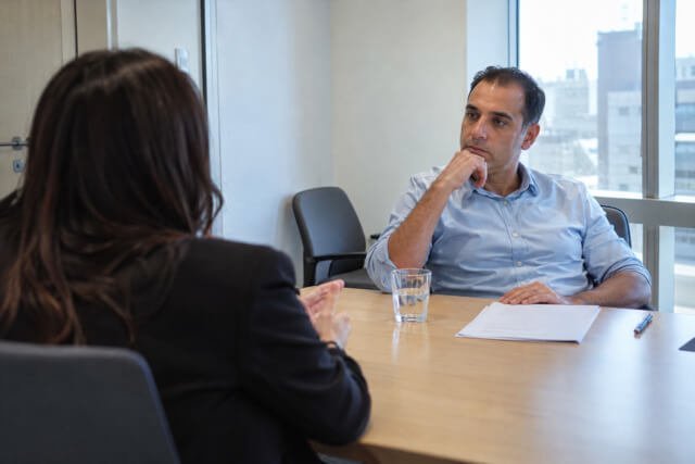 Homem observa uma candidata durante uma entrevista de contratação em uma sala de reunião. Imagem utilizada para ilustrar artigo O seu instinto pode ser medo antigo respondendo por você? de Homero Mônaco Psicanalista especializado em neurociências e comportamento.