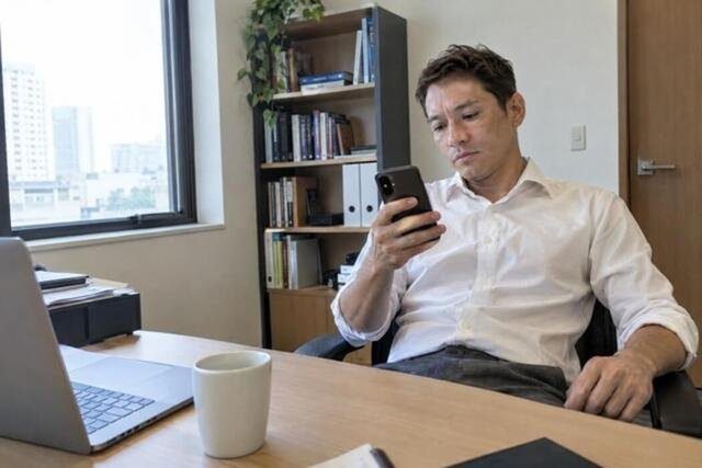 Homem sentado à mesa de um escritório particular olha para uma mensagem positiva de um cliente no celular e se prepara para encaminhá-la ao sócio. A cena representa a satisfação breve com o reconhecimento profissional e a busca imediata por uma nova confirmação. Imagem utilizada para ilustrar o artigo “Você realmente está atrás do seu reconhecimento profissional ou no fundo precisa de outra coisa?” escrito por Homero Mônaco Psicanalista especializado em neurociências e comportamento.